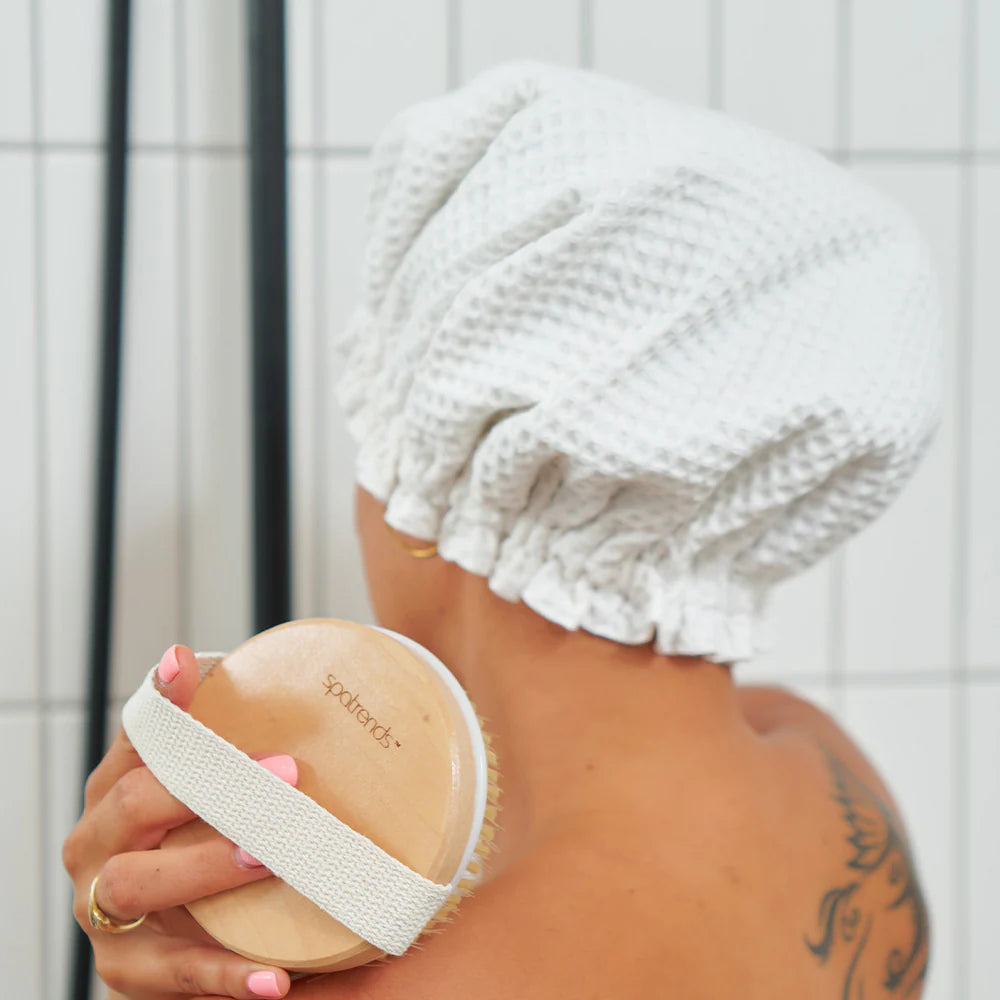 A woman facing away from the camera and body brushing her shoulder in the shower whilst wearing the Annabel Trends waffle white shower cap.