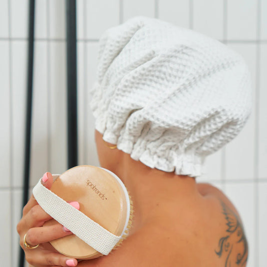 A woman facing away from the camera and body brushing her shoulder in the shower whilst wearing the Annabel Trends waffle white shower cap.