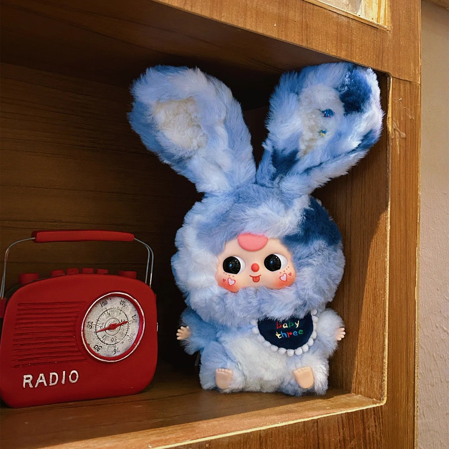 Blue plush toy with bunny ears on a wooden shelf next to a red radio.