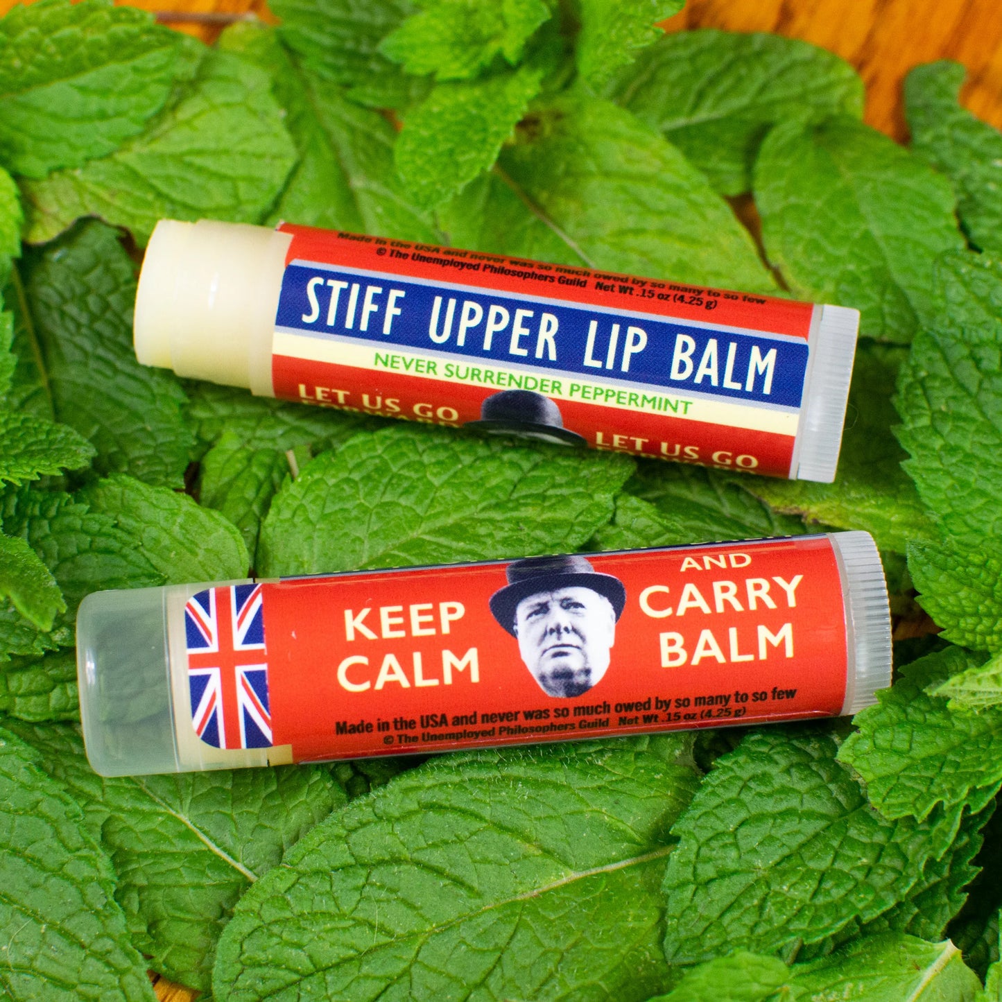 Two lip balms on a bed of green leaves, one labeled 'Stiff Upper Lip Balm' and the other 'Keep Calm and Carry Balm'.