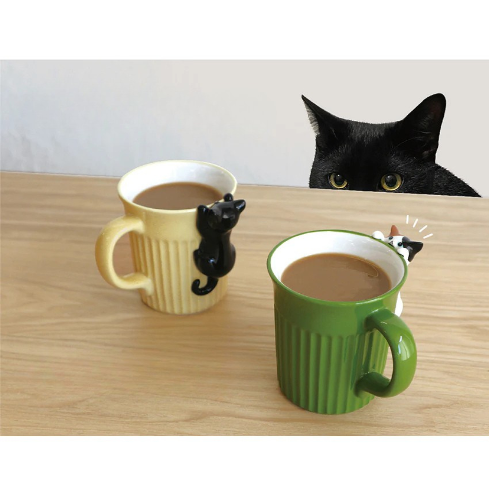Two mugs with cat designs on a table, one yellow and one green, with a black cat peeking from behind.