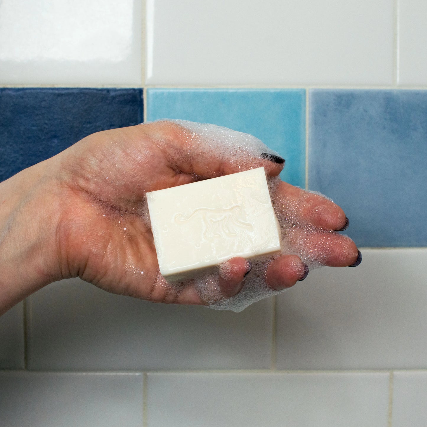A hand displaying a bar of soap with suds with a blue tiled wall in the background.