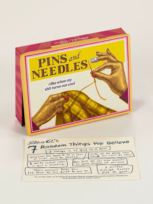 A yellow pocket sized tin box from Blue Q featuring a Pins and Needles title. The image displays a lady's hands sewing a thread across a yellow tartan fabric. Below is a small note stating the beliefs of Blue Q mission statement.