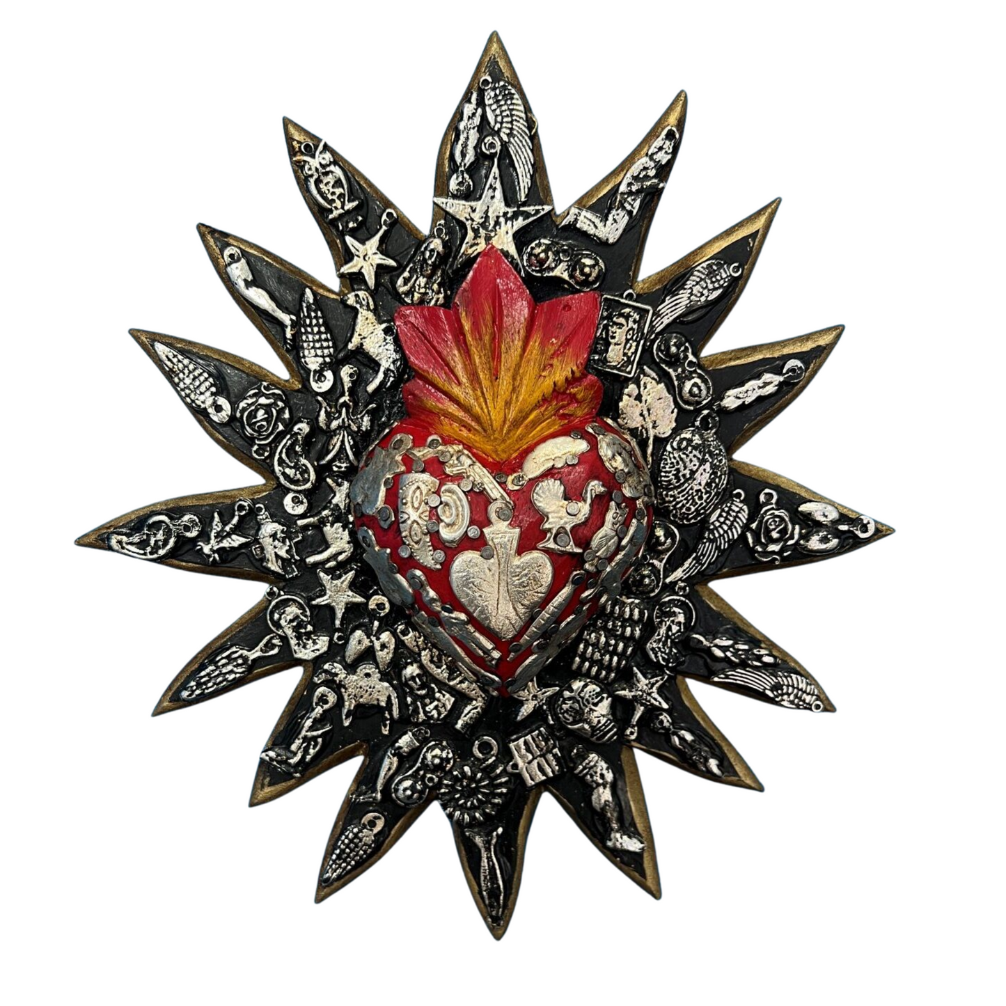 Wooden Mexican wall decor- Black Sun shape with a central red heart motif. The ornament is covered in charms which have a spiritual meaning. The decor piece is on a white background.