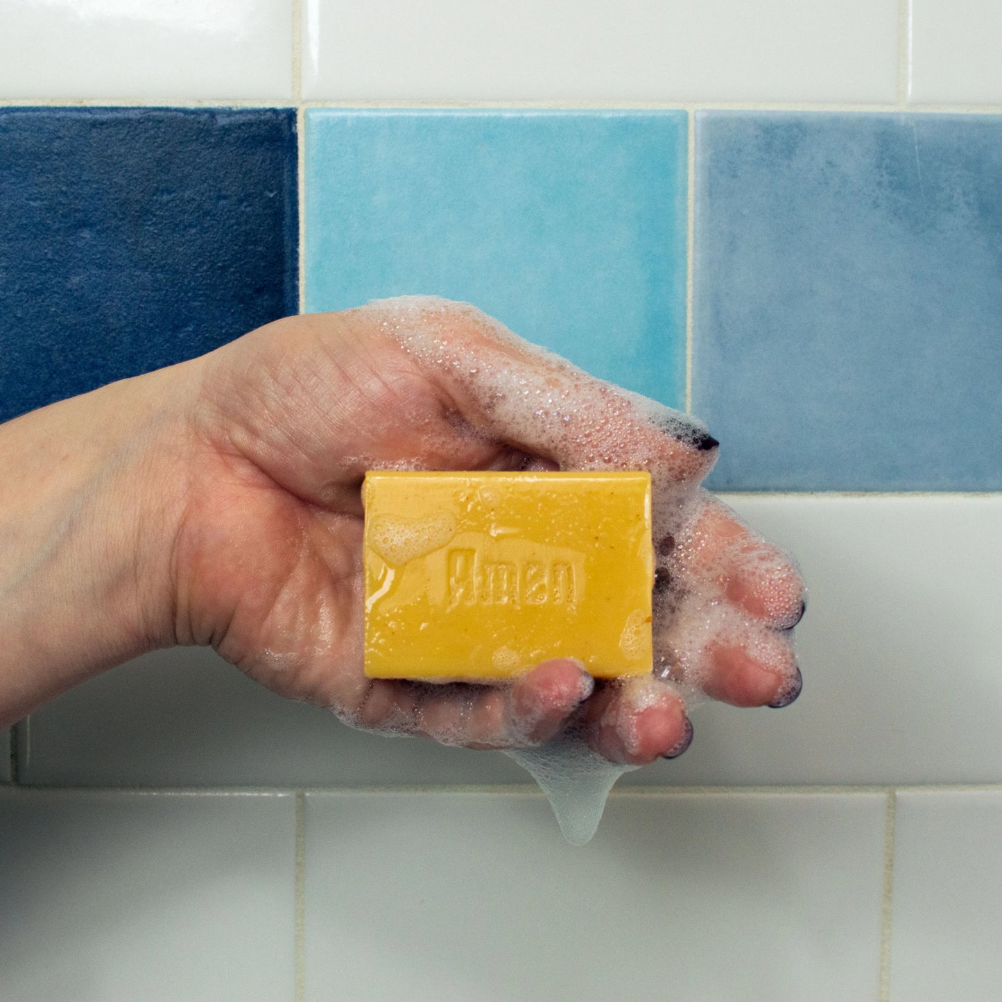 Hand holding a yellow bar of soap with 'Almen' branding against a tiled bathroom wall.
