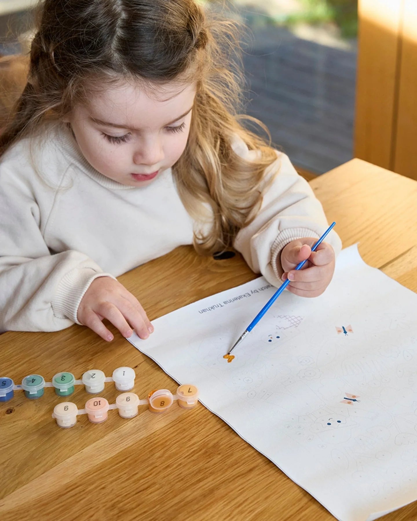 Child painting on a piece of paper with a brush at a wooden table.