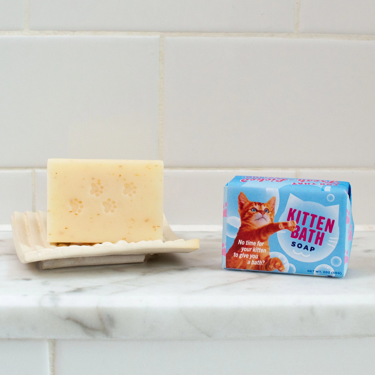 a bar of soap on a soap dish next to a blue packaged bar of kitten hand soap. The background is a white marble ledge and tiled wall.