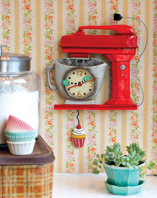 Red stand mixer with a clock face on a floral wallpapered wall.
