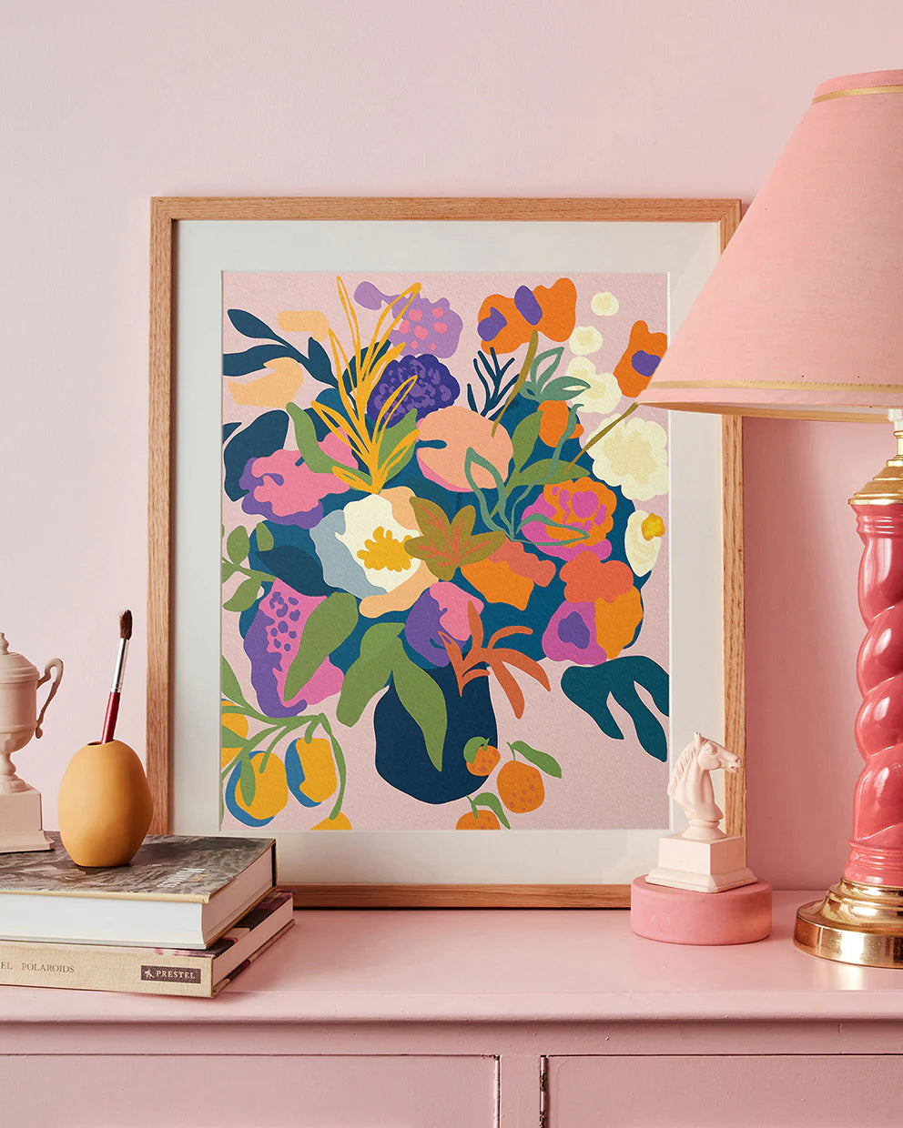 Framed colorful floral "In Bloom"artwork on a pink wall with decorative items.