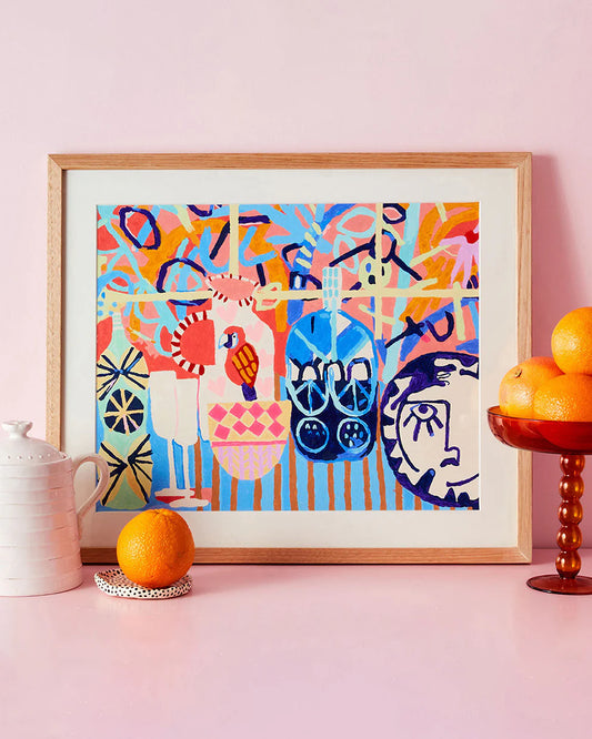 Framed colorful abstract art piece on a pink wall with oranges and a teapot.