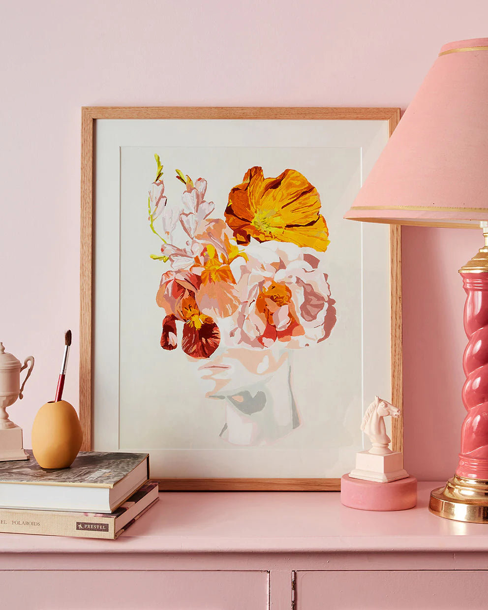 Floral artwork on a pink shelf with a lamp and books