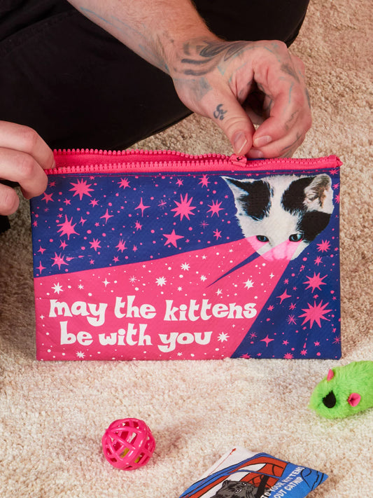 A man's hand with a tattoo, zippering the may the kittens be with pouch pouch . He is sitting on a beige carpet surrounded by cat toys.