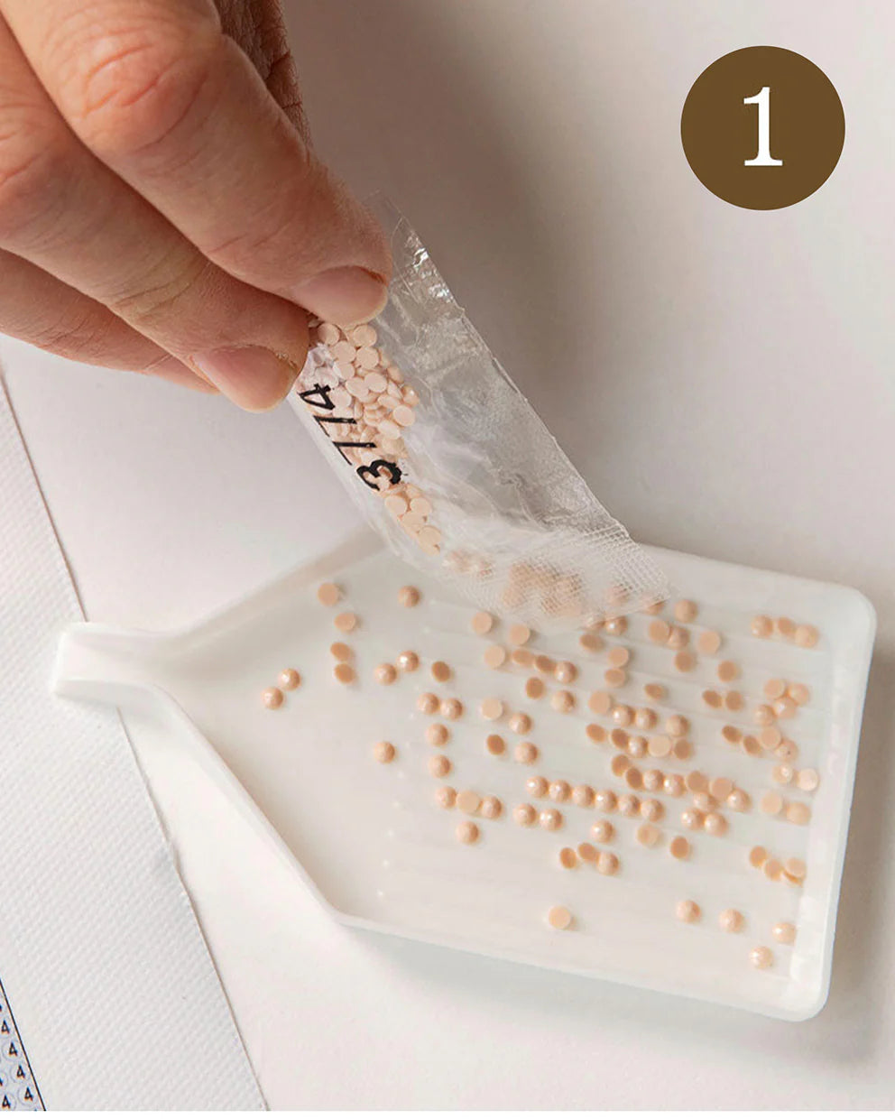 Hand pouring small beige beads from a clear plastic bag onto a white surface.