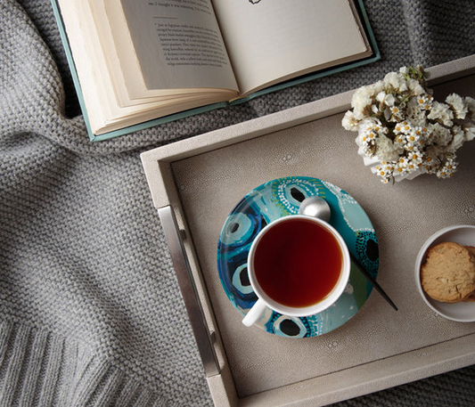 Tea cup and saucer set by May Wokka Chapman placed on a neutral tray. To the left there is a small floral arrangement in a vase and a couple of biscuits to o with the tea. An open paged book layson a grey blanket.