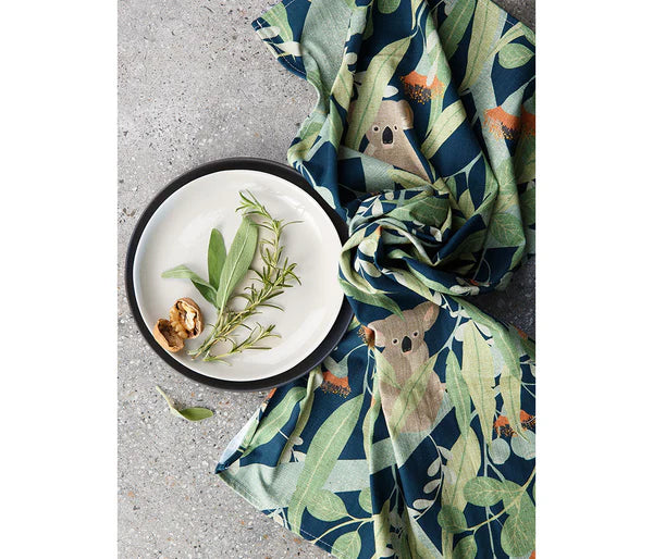 Koalas print cotton tea towel placed near a plate with a walnut shell and rosemary placed on a background of concrete.