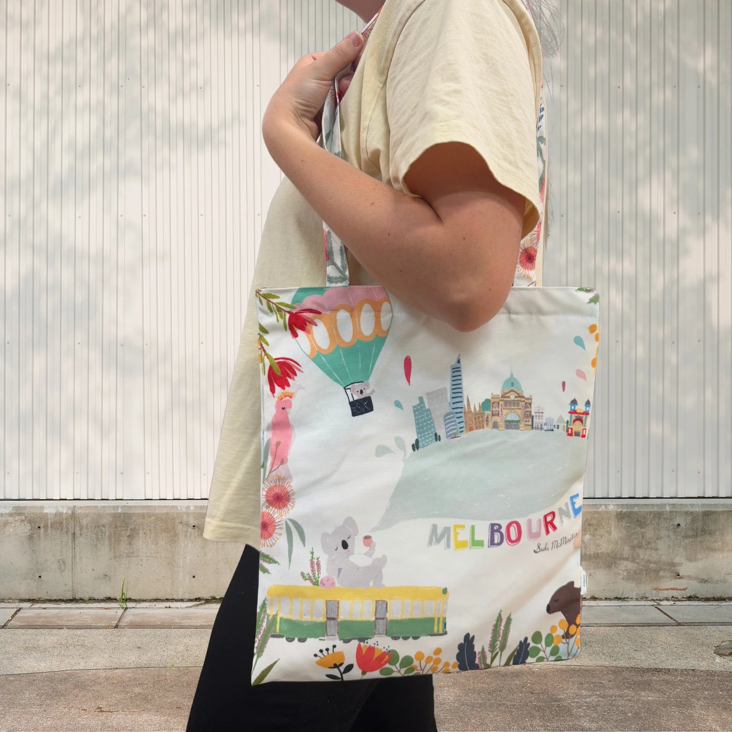 Person holding a Suki McMaster tote bag with a colorful design featuring Melbourne landmarks and animals.