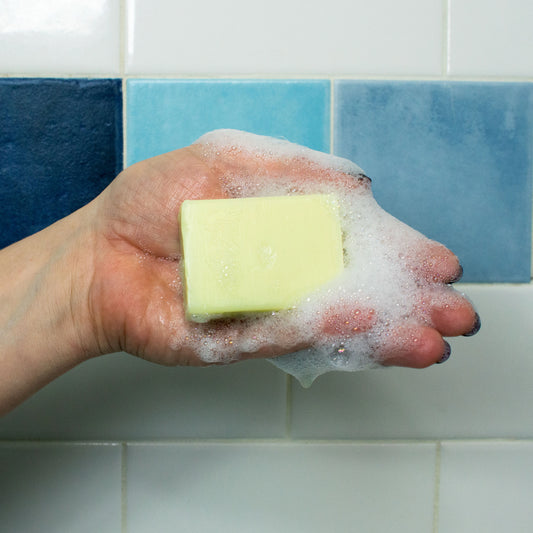 Hand holding a bar of soap with soap suds against a tiled bathroom wall.