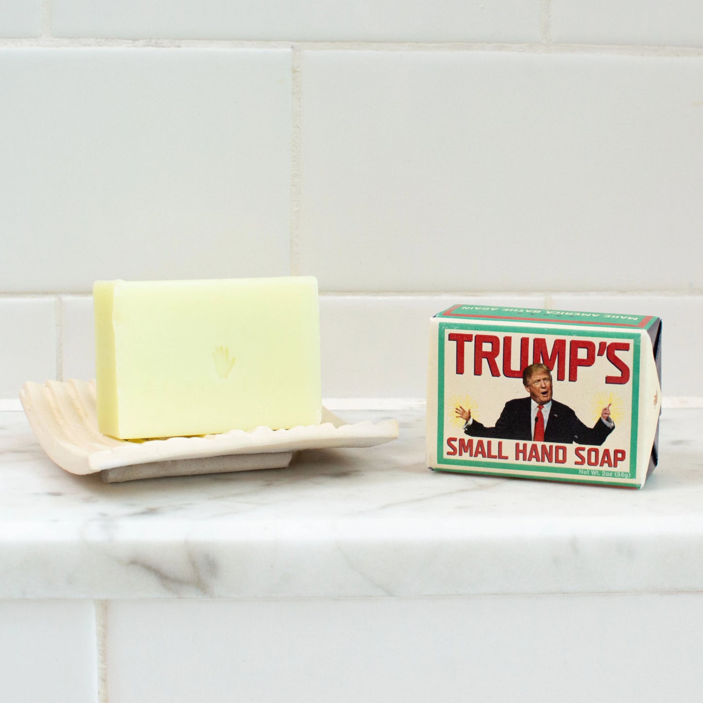 Bar of soap on a soap dish next to a box labeled 'TRUMP'S SMALL HAND SOAP' on a tiled wall background.