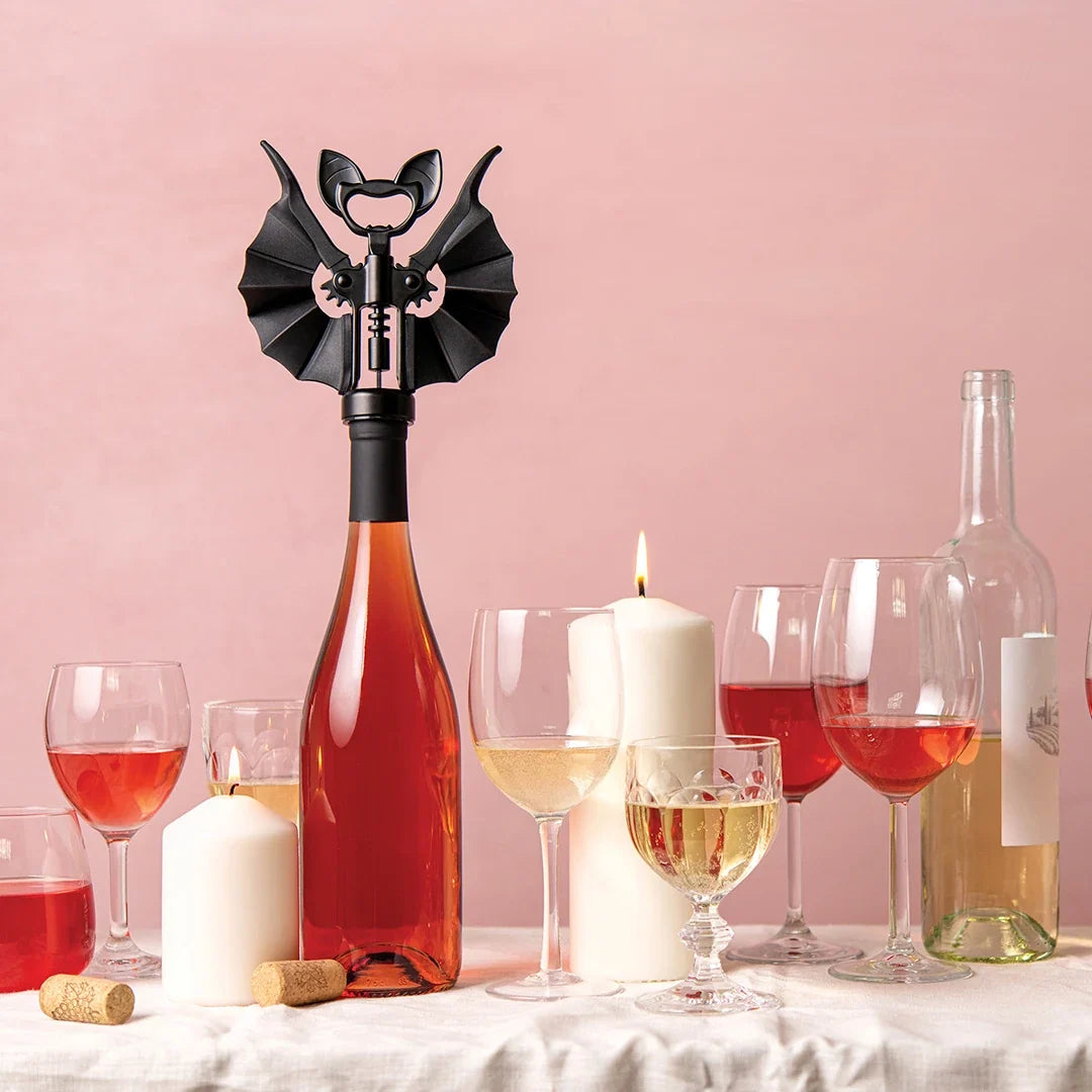 A bat shaped bottle opener on a bottle of wine with a setting of white wine bottles and glasses filled. Two lit white candles and two corks in the foreground. The background is pink.