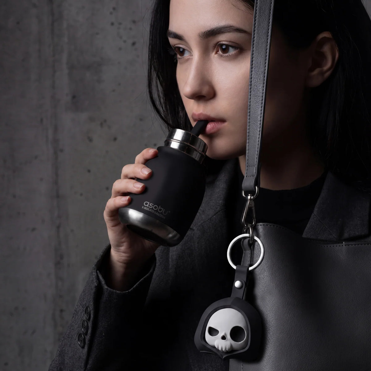 A female dressed in black taking a sip from a black mini drink bottle, an a strap hanging the bottle op that has a skeleton face. The background is grey.