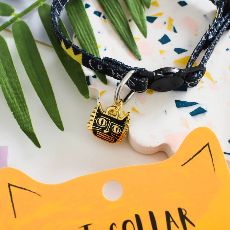 Black and gold cat-shaped charm on a black cord with a yellow tag labeled 'Cat Collar' on a colorful background.