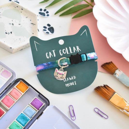 Cat collar with colorful tie-dye design and cat charm on a green card with text, surrounded by art supplies.