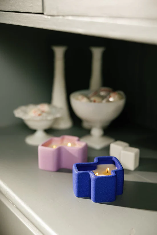 Two candles in colorful holders on a shelf with decorative items in the background.