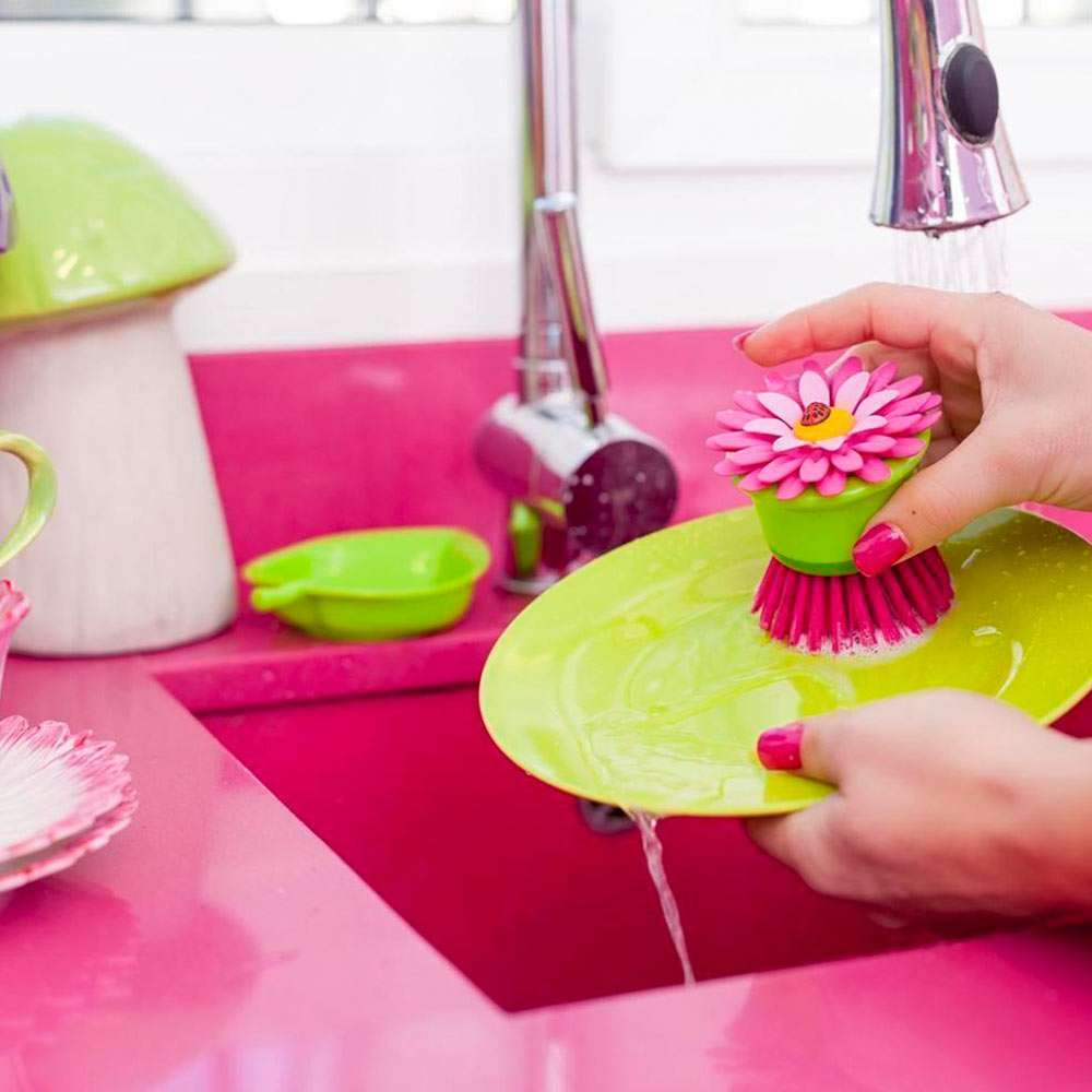 Person using a pink flower-shaped scrubber to clean a green plate in a pink sink.