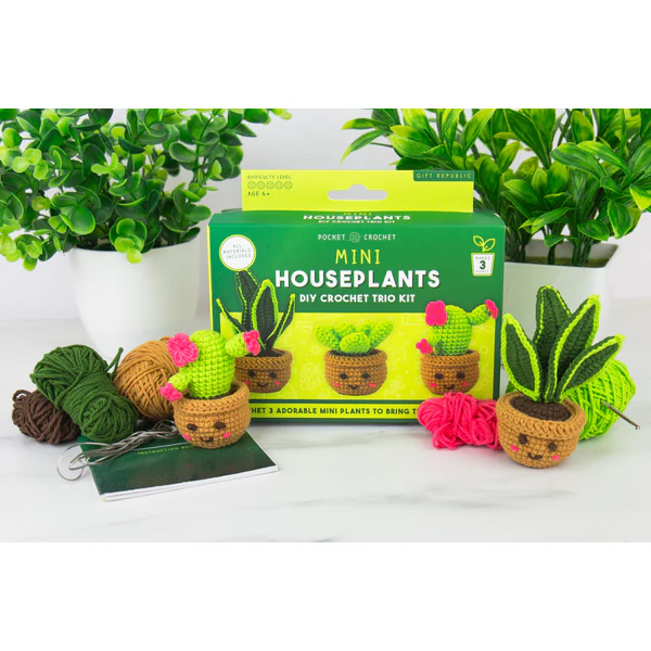 A crochet kit box labeled 'Pocket Houseplants Mini Houseplants DIY Crochet Kit' displayed on a white surface with small crocheted cactus and succulent plants, yarn balls, and greenery in the background.