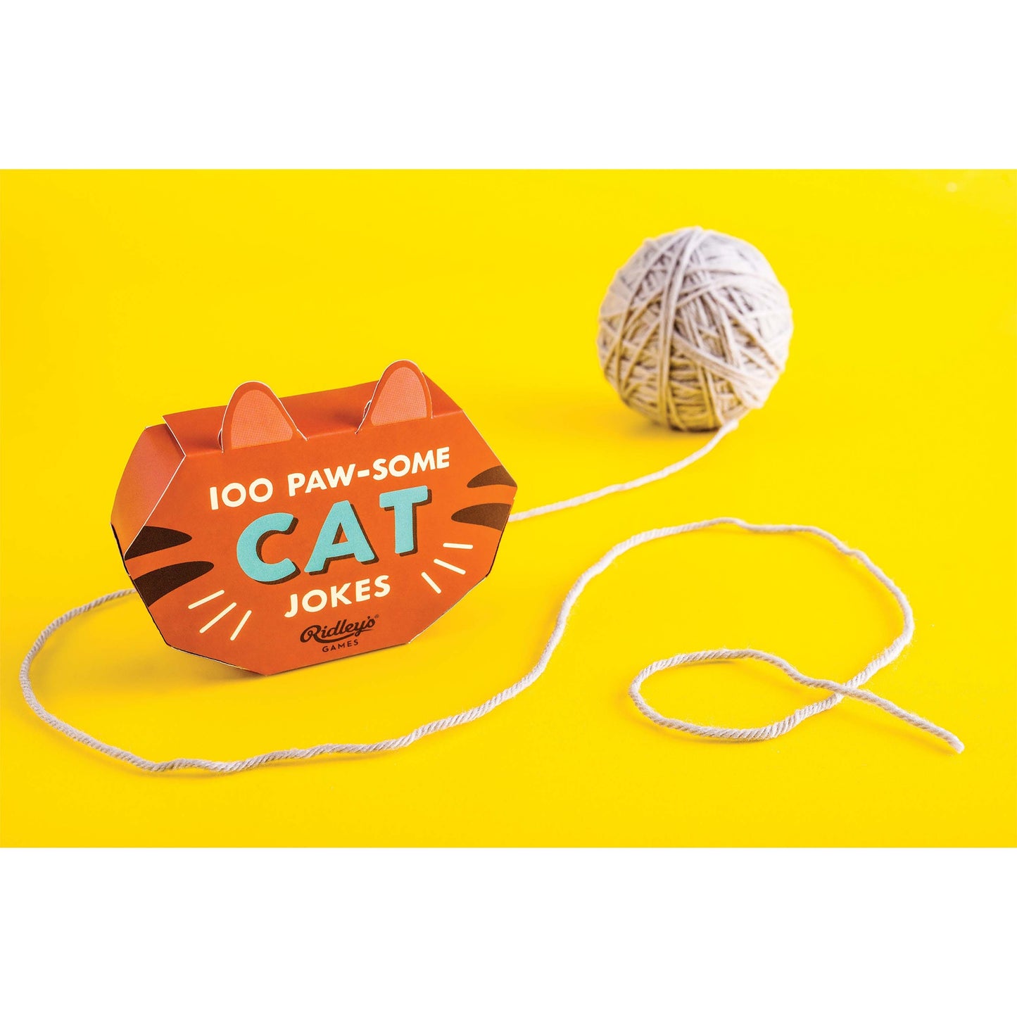 A ball of string next to a box of '100 PAW-SOME CAT JOKES' on a yellow background
