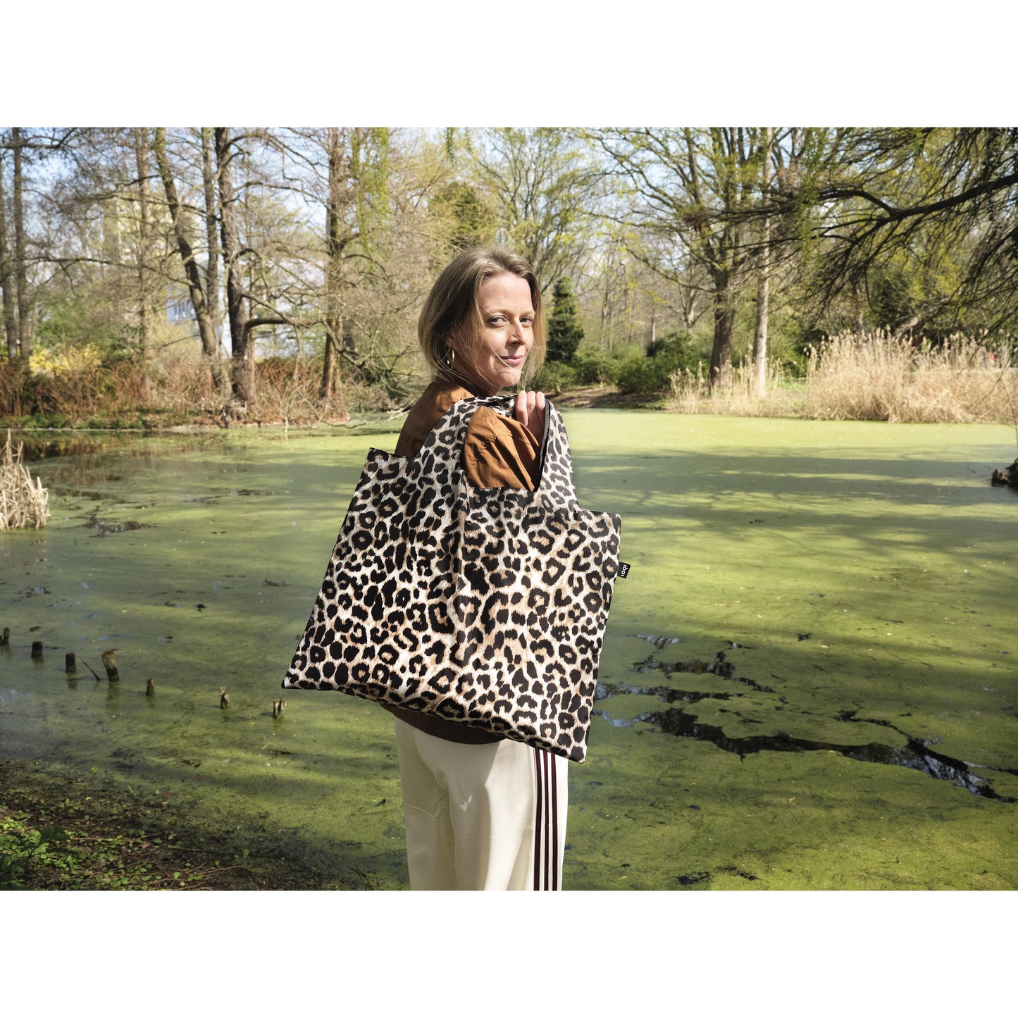 A woman overlooking a green wooded area and pond, looking back whilst holding Leopard shopping bag over her shoulder.