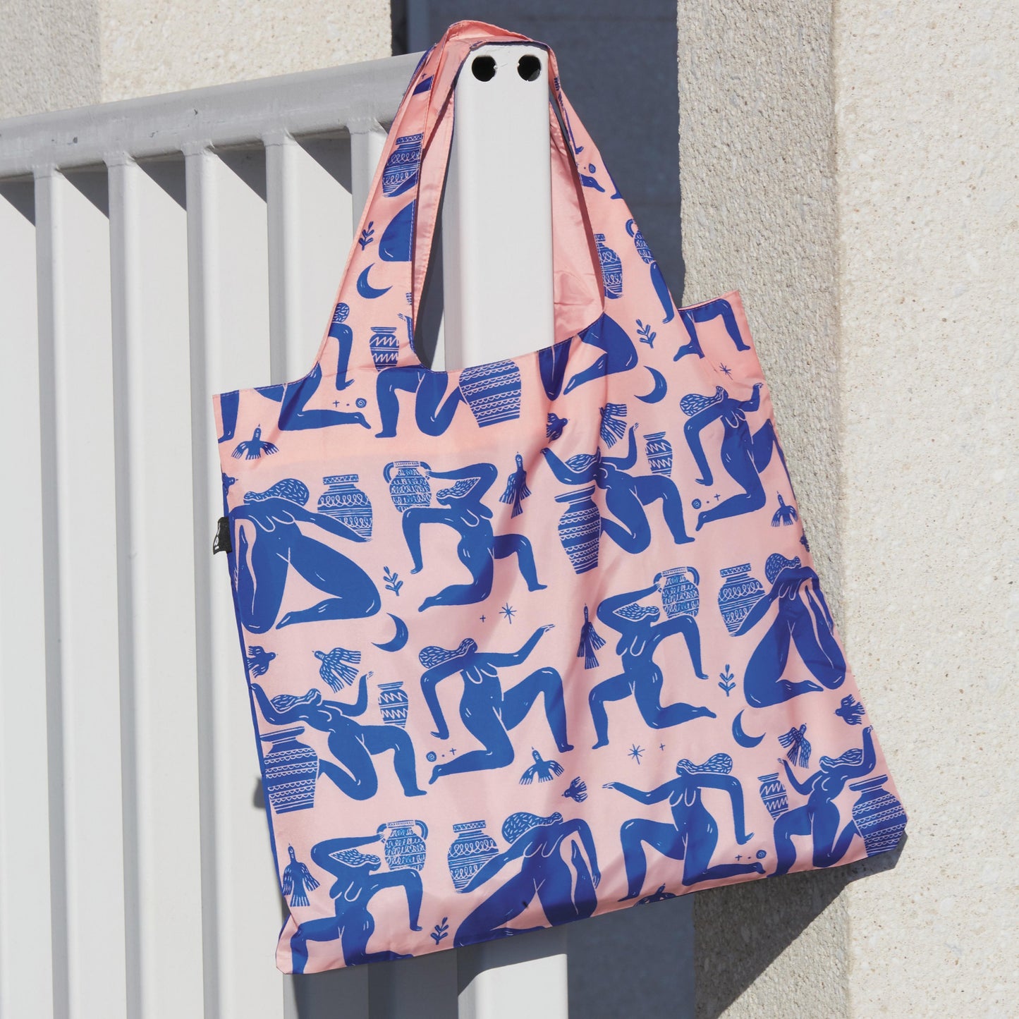 Reusable shopping bag with blue figure pattern on pink background, hanging on a railing.