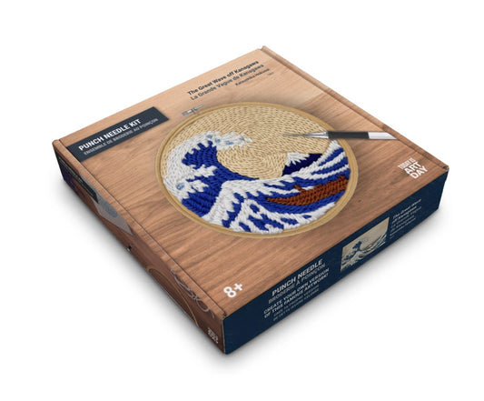 A boxed punch needle kit shown at an angle on a white background. The box is wood-toned with a circular preview of a blue, white, and tan wave design, plus a small paintbrush illustration.