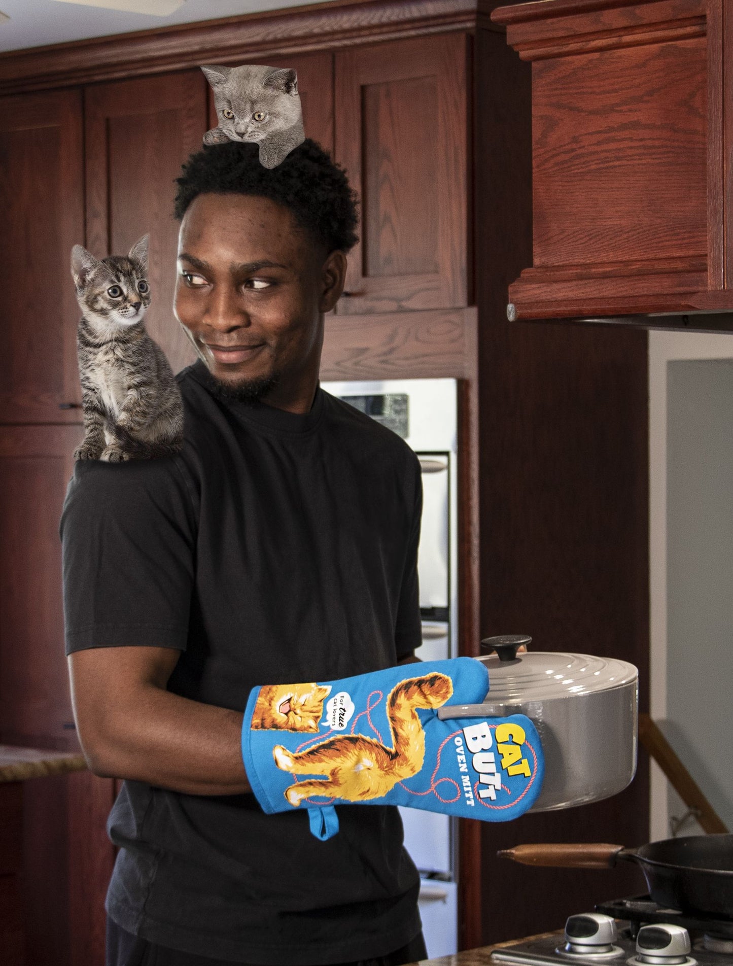 A photograph of a man in a kitchen holding a casserole dish and wearing the BLUEQ Oven glove- Cat Butt. He balances two kittens whilst smiling.