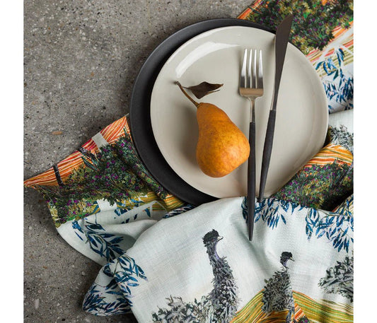 A Tea towel - Australian indigenous art by Brigid (Emu) placed underneath a plate of cutlery and a pear. The background is  concrete  - Gizmo Gifts