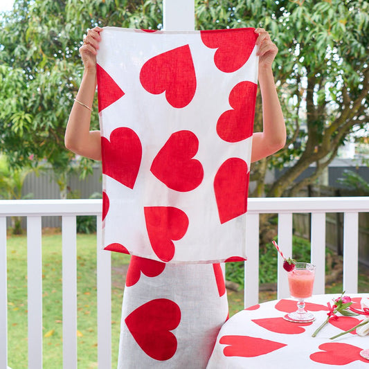 A woman holding up a printed teatowel - Love Hearts (Linen) ,wearing an apron and standing near a tablecloth of the same design- The background is a lush green backyard.Gizmo Gifts