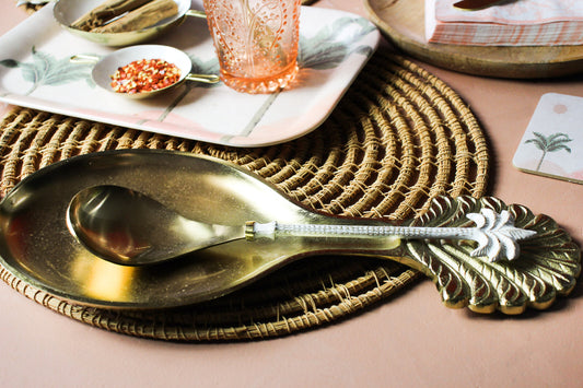 Decorative spoon on a woven placemat with a pink glass and napkin in the background.