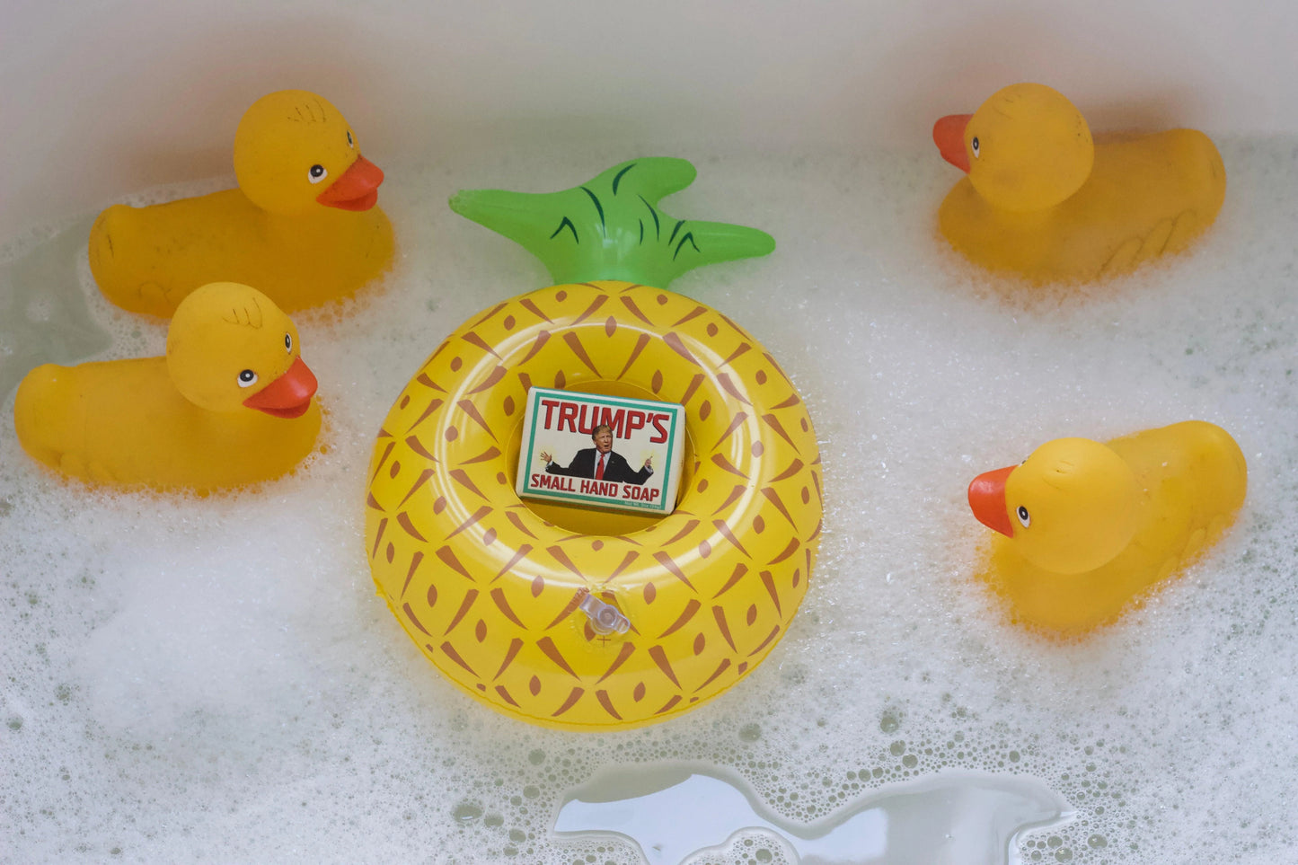 Yellow rubber duckies and a pineapple-shaped soap with 'TRUMP'S' logo in a bathtub.
