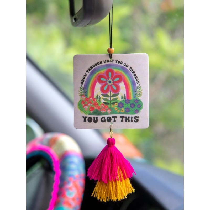A square car air freshener or hanging ornament with a colorful rainbow flower illustration and the words 'CARE THROUGH WHAT YOU GO THROUGH' and 'YOU GOT THIS,' suspended by a black string in a car interior. A bright pink and yellow tassel hangs below.