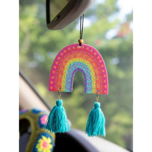 A colorful rainbow-shaped car hanging ornament with two turquoise tassels, shown hanging from a rearview mirror in a car with a blurred green outdoor background.