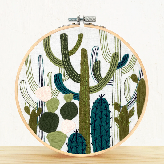 Completed Cactus Garden embroidery hoop featuring hand-stitched cacti and succulents in soft green and earthy tones on cotton fabric, framed in a 6-inch bamboo hoop." White background and hoop placed on a pine shelf.