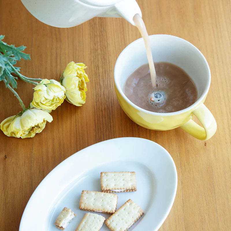 Tea being poured into a yellow mug on a wooden table with cookies and flowers.