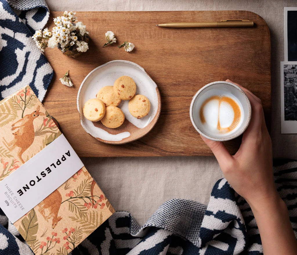 An outstretched hand holding a coffee and a table  spread containing a plate of biscuits and an Applestone package in the foreground.