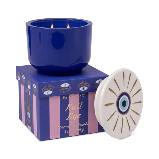 Blue candle in a decorative box with ' Evil Eye' design, next to a small round object with similar design.