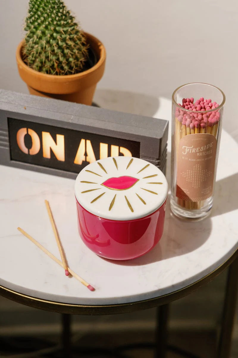 Pink candle with white lid on a table next to matches and a small plant.