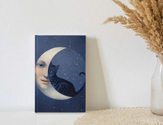 A navy blue journal featuring the art of Catrin Welz-Stein. The artwork shows a moon face with a black cat sitting within its surface.