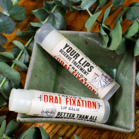 Two lip balm tubes labeled 'Oral Fixation' on a wooden surface with green leaves.