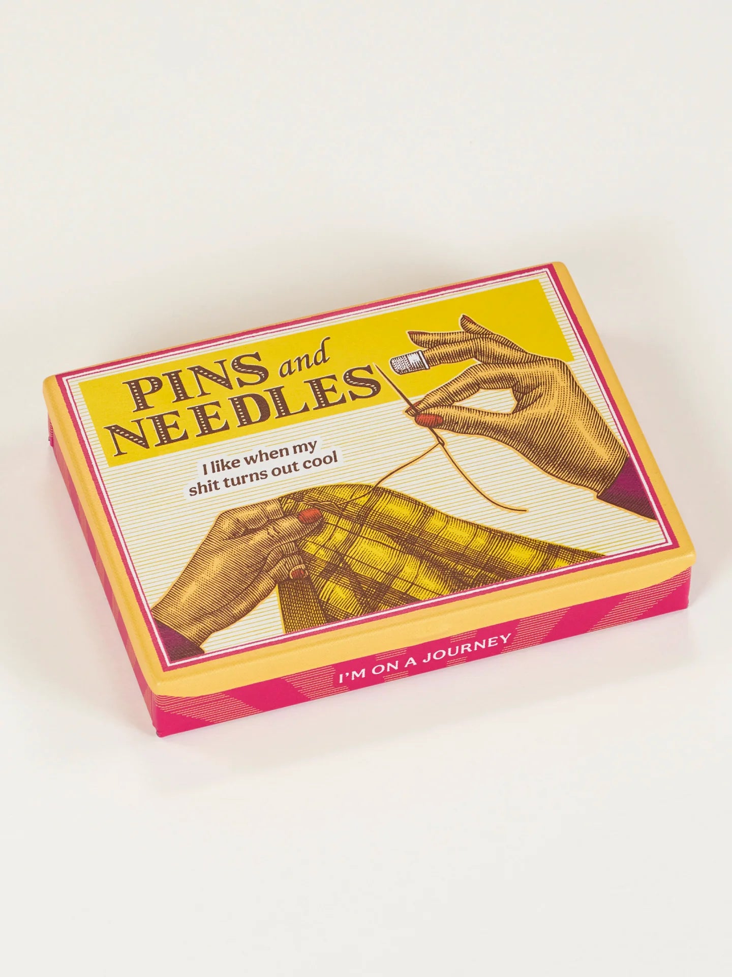 A yellow pocket sized tin box from Blue Q featuring a Pins and Needles title. The image displays a lady's hands sewing a thread across a yellow tartan fabric. it sits on a white background,flatlay.