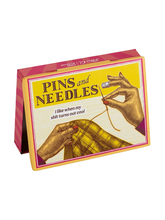 A yellow pocket sized tin box from Blue Q featuring a Pins and Needles title. The image displays a lady's hands sewing a thread across a yellow tartan fabric.