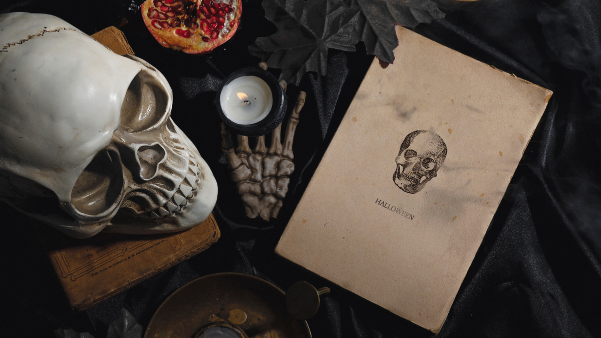A spooky black background with a white skull , a pomegranate, a skeletal hand holding a lit candle and printed sheet of paper with text that reads: Halloween.